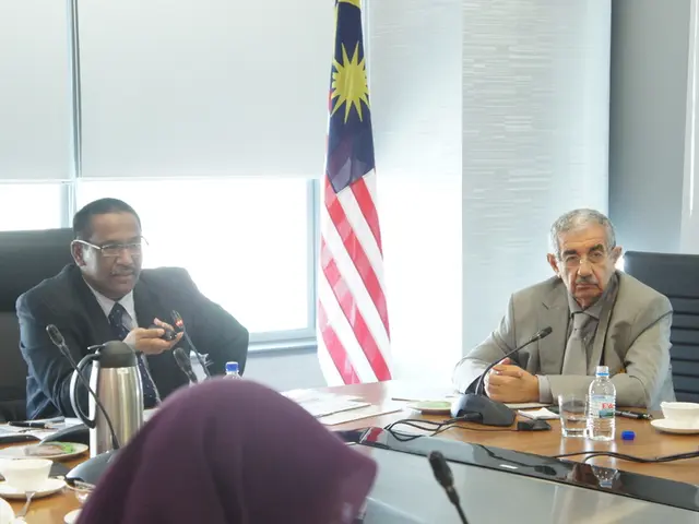 In the image there are men in suits and two men women in burqa sitting around table with water...