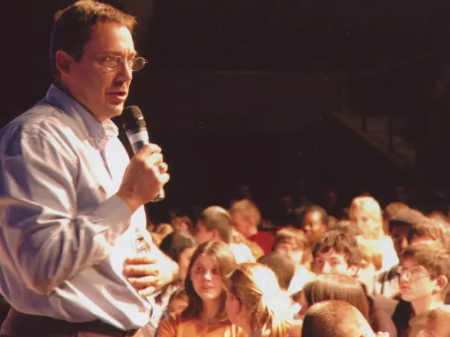 In this picture there is a man wearing a blue color shirt is giving a speech. Beside there is a...