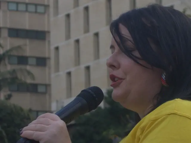 A lady in yellow dress wearing a watch is holding mic and talking. In the background there is a...
